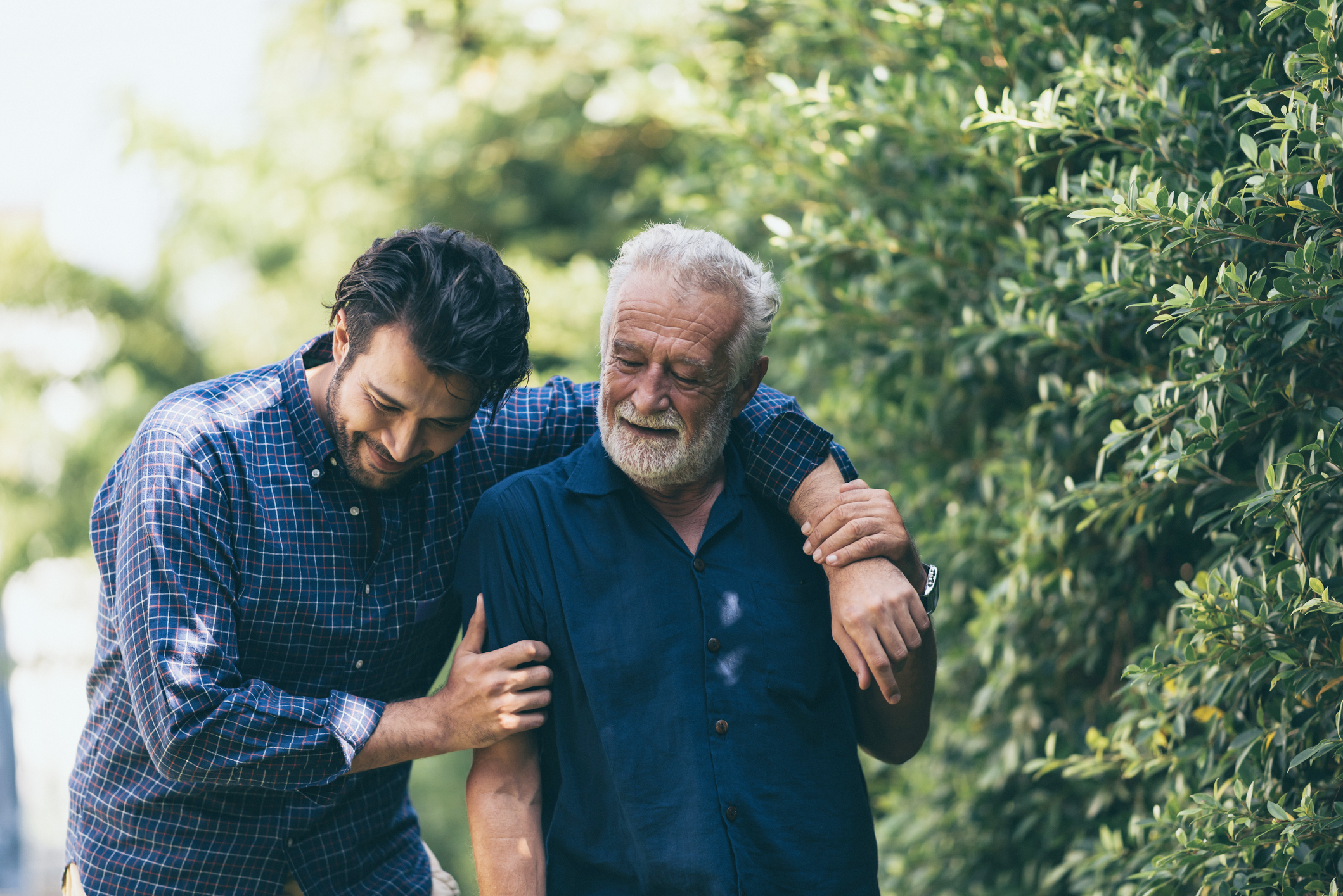 The old man and his son are walking in the park. A man hugs his elderly father. They are happy and smiling